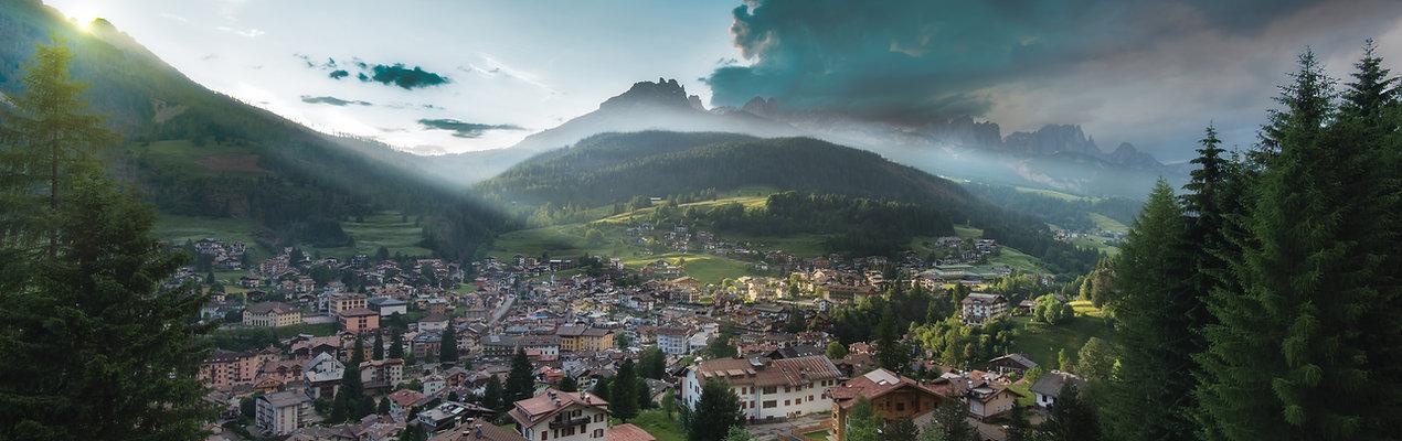 Panorama montano con villaggio, colline verdi e luce solare che filtra tra le nuvole.